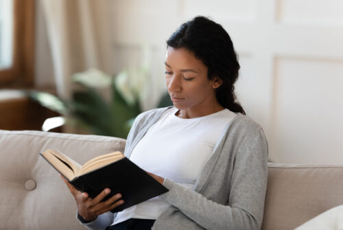 Focused African American female sit relax on sofa in living room reading interesting book, concentrated biracial young woman rest on couch at home enjoying literature novel, spend weekend indoors